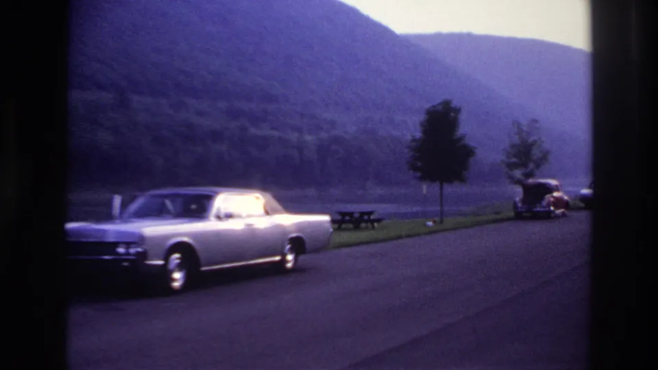 1967 Ottawa-area 8mm frame with the silver sedan drifting toward the left edge, a lakeside road, picnic table, trees and hills beyond.