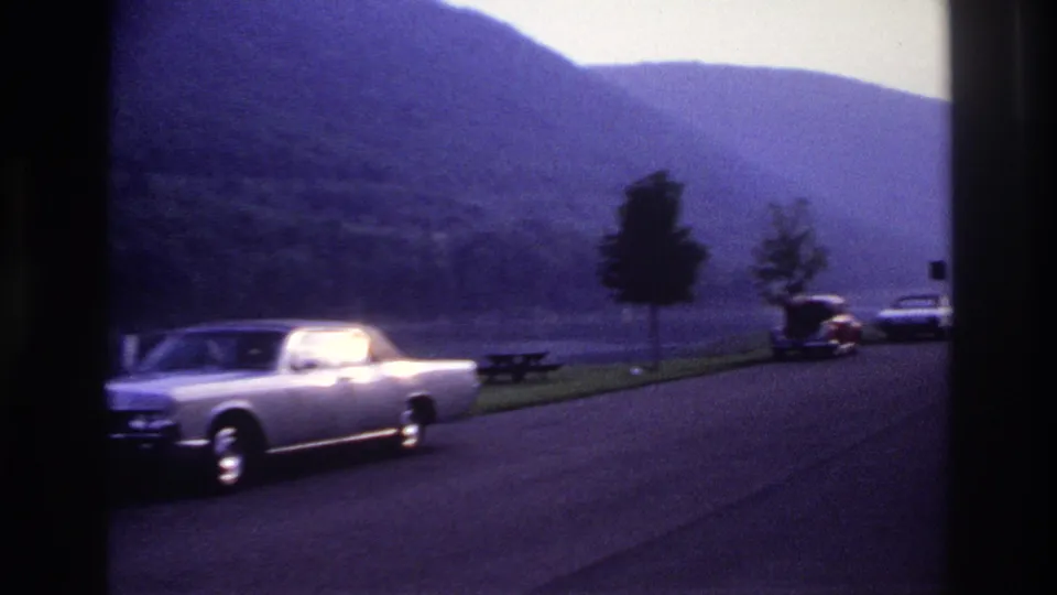 1967 Ottawa-area home-movie frame showing the silver sedan at left with a lakeside park road, picnic table, trees, wooded hills and distant cars.
