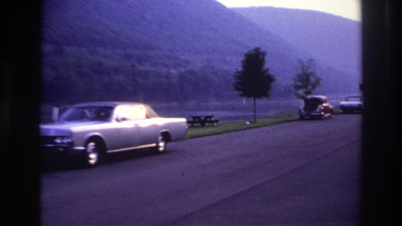 Silver convertible on a road near parked cars, picnic tables, lake, and hills.