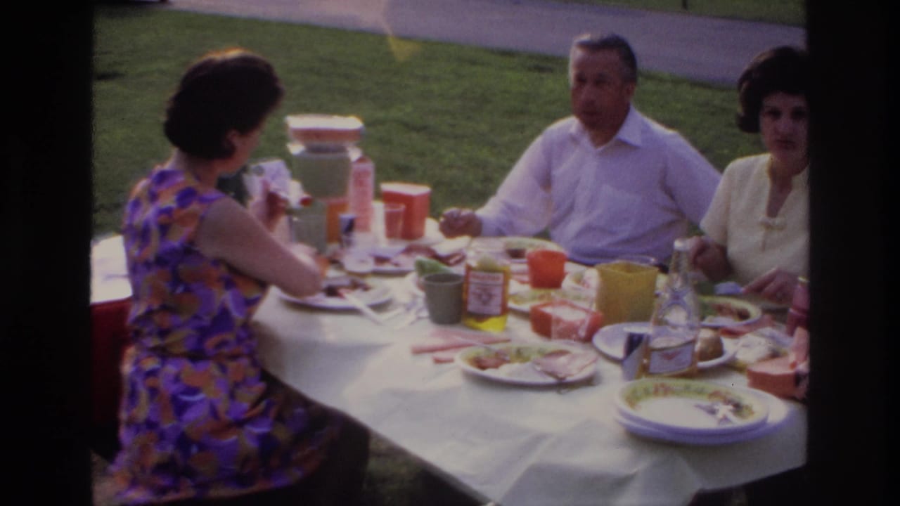 Three adults sit at a white-clothed picnic table on grass with plates, cups, condiments, and food containers.