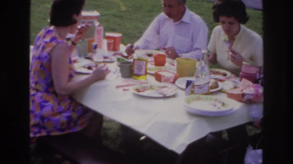 1967 Ottawa picnic frame showing adults mid-meal at a white-clothed table with cups, plates, food containers, and grass behind them.