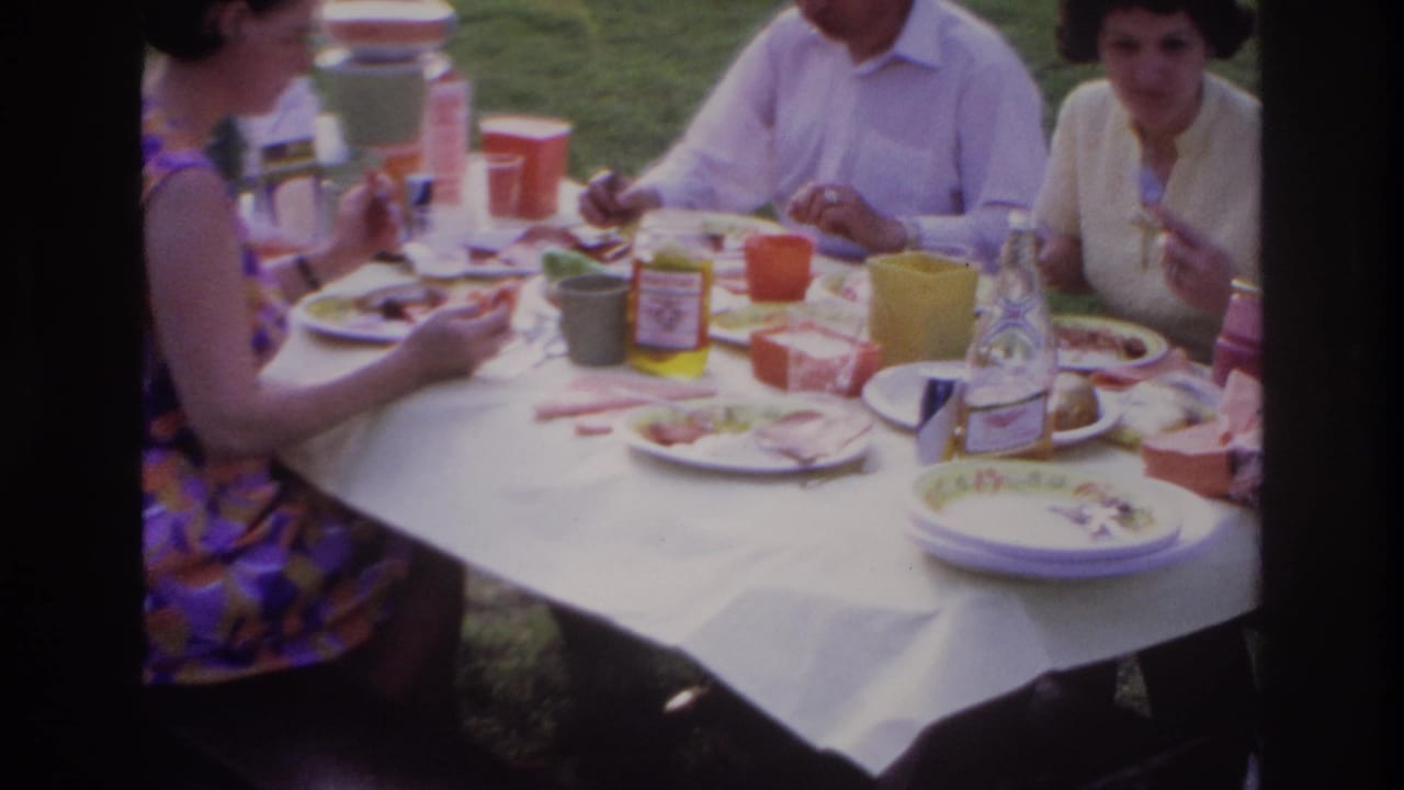 1967 Ottawa home-movie frame showing adults eating at a park picnic table with colorful cups, plates, cold cuts, and a pickle jar.