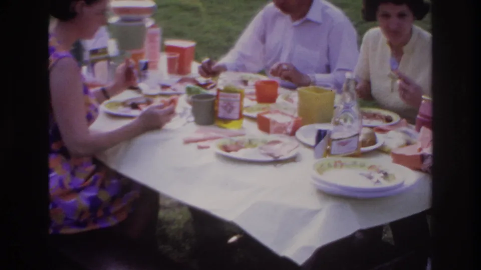 1967 Ottawa home-movie frame showing adults eating at a park picnic table with colorful cups, plates, cold cuts, and a pickle jar.