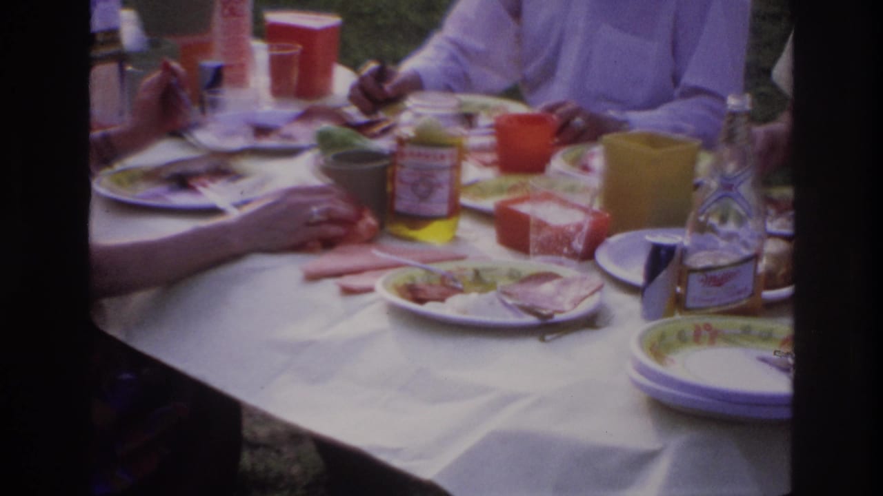 Close view of picnic table items with adults' hands, cups, plates, and a Magyar jar.