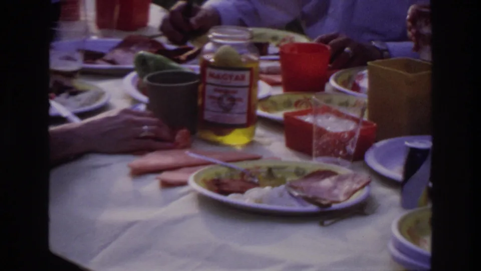 Close 8mm frame from the Ottawa picnic showing plates of food, plastic cups, napkins, and a Magyar jar on the table.