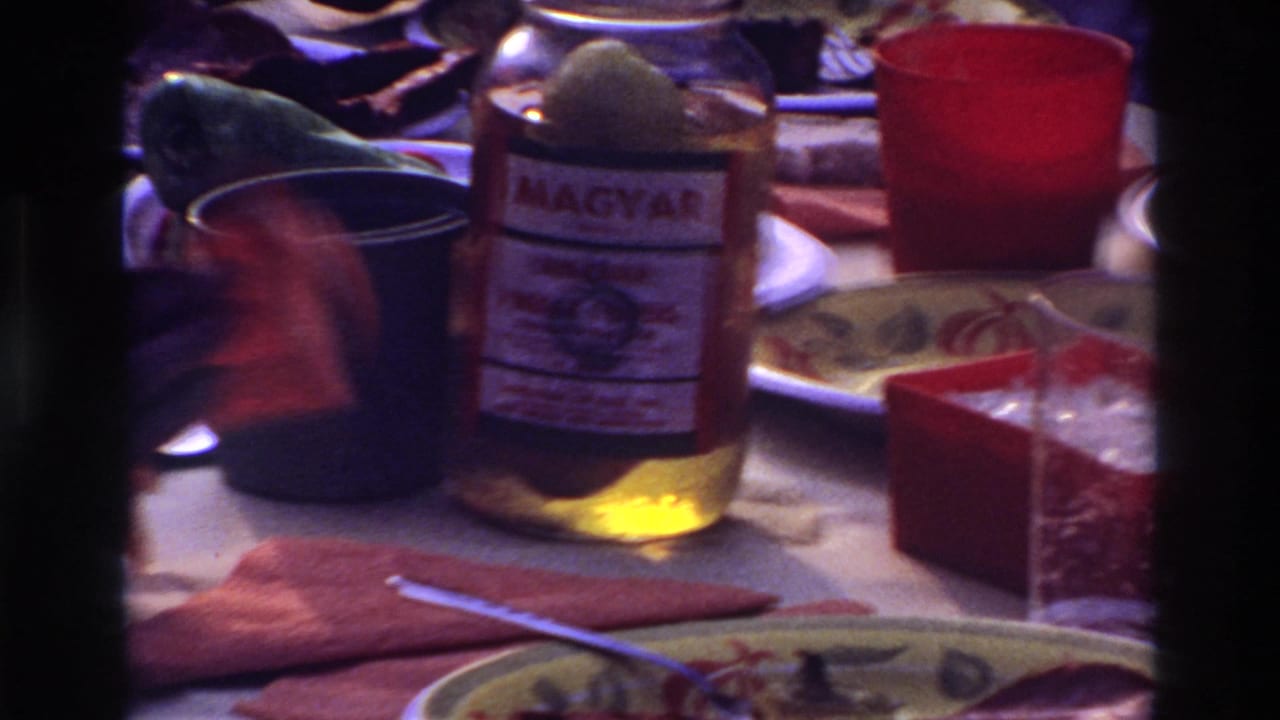 1967 Ottawa home-movie frame showing a crowded lunch table with a Magyar-labeled jar, red cup, dark mug, plates, and partial hands.
