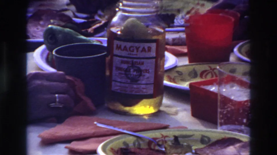 1967 Ottawa home-movie frame showing a close view of the Magyar-labeled jar, dark mug, red cup, plates, napkin, and a hand.