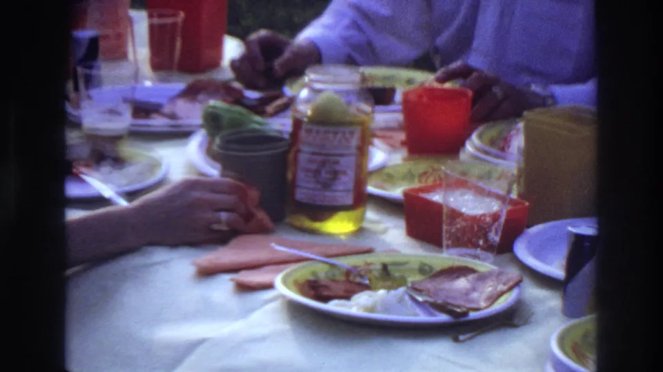 1967 Ottawa home-movie frame showing a wider lunch table with a Magyar-labeled jar, cups, plates, hands, and a partial person in a light shirt.