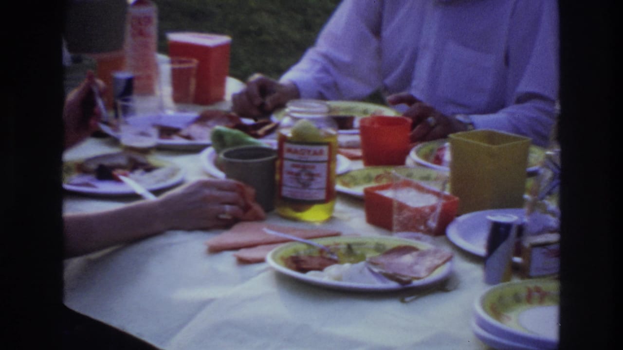 1967 Ottawa home-movie frame of adults eating outdoors at a white-covered picnic table with colorful dishes and condiments.