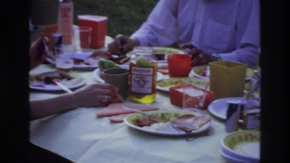 1967 Ottawa home-movie frame of adults eating outdoors at a white-covered picnic table with colorful dishes and condiments.