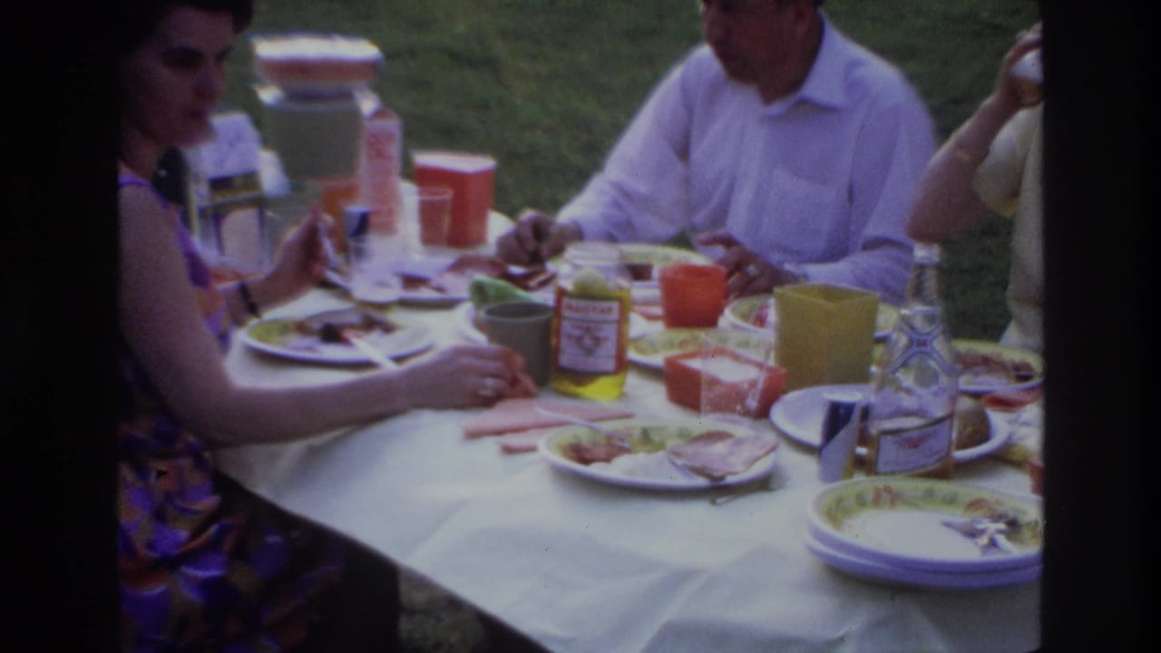 Adults sit at an outdoor table with plates, cups, a drink bottle, and condiment jars visible.