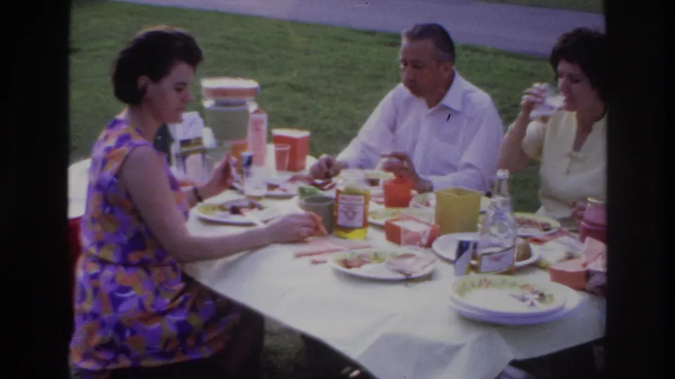 1967 Ottawa frame showing adults in summer clothing eating at a picnic table, including a woman in a purple-orange patterned dress.
