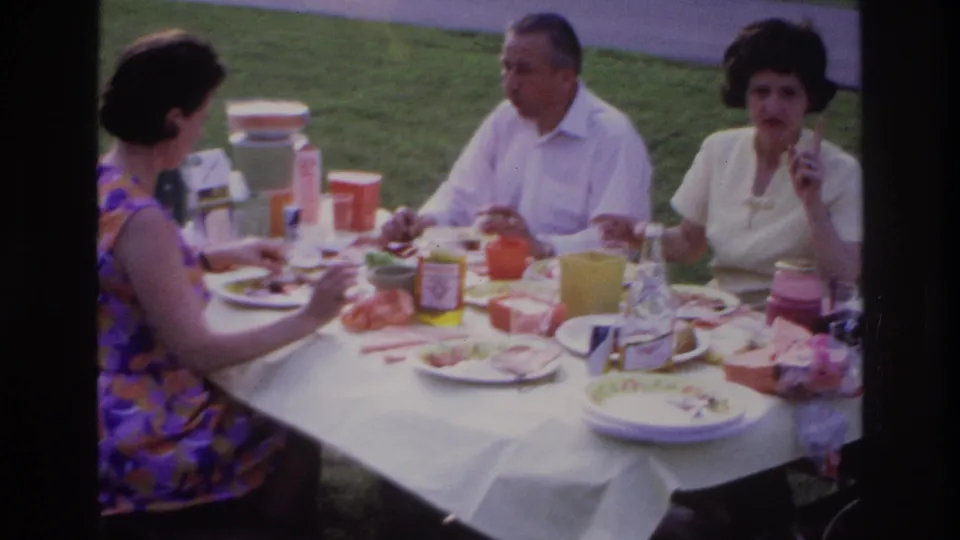 Later 1967 Ottawa picnic frame with adults seated around a white-clothed table, eating and drinking on a grassy lawn.