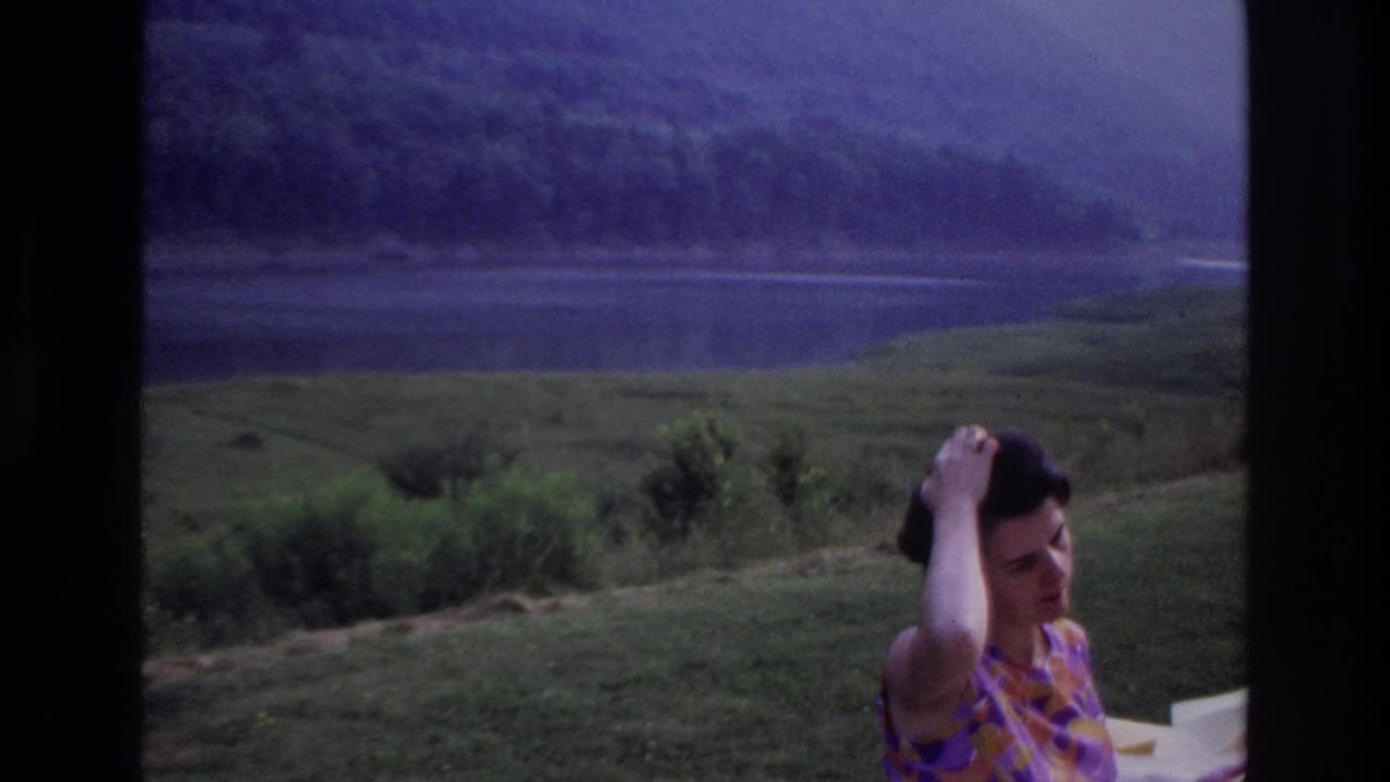 1967 Ottawa-area home movie frame of a woman in a patterned dress adjusting her hair near a lakeside picnic table.