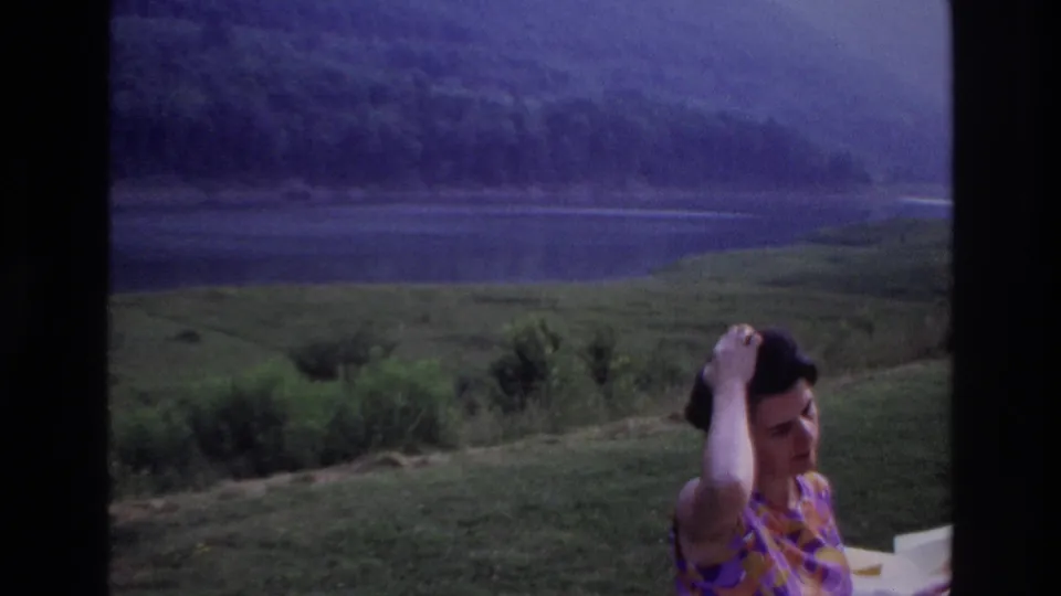 1967 Ottawa-area home movie frame of a woman in a patterned dress adjusting her hair near a lakeside picnic table.