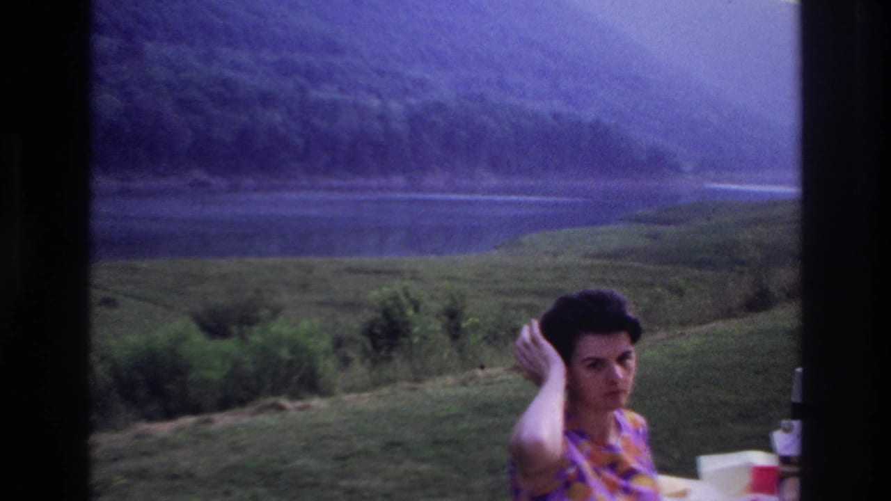 Woman looks toward the camera while touching her hair near a lakeside picnic table.