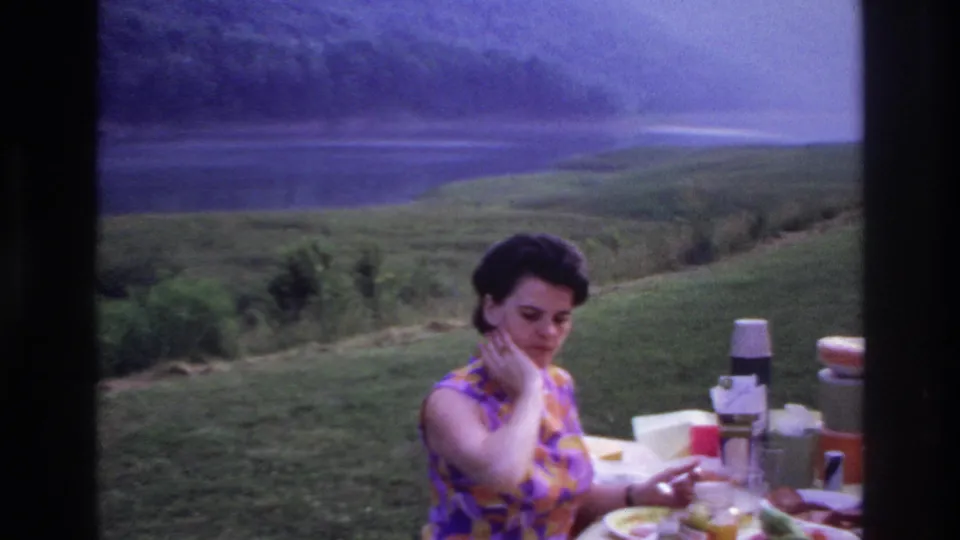 1967 home movie frame showing a woman at a white-clothed picnic table with food and drink containers before a lakeside landscape.
