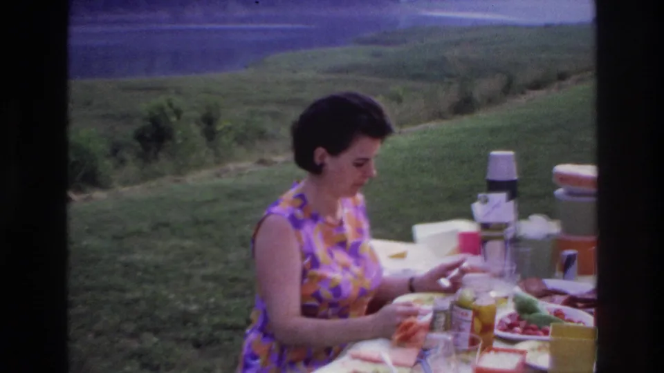 Woman in a patterned dress looks down at a lakeside picnic table set with food, cups, jars, and a thermos in 1967 Ottawa footage.