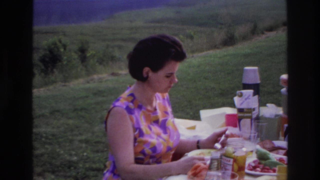 1967 Ottawa home-movie frame of a woman eating at an outdoor table with food and picnic items on a grassy hillside.