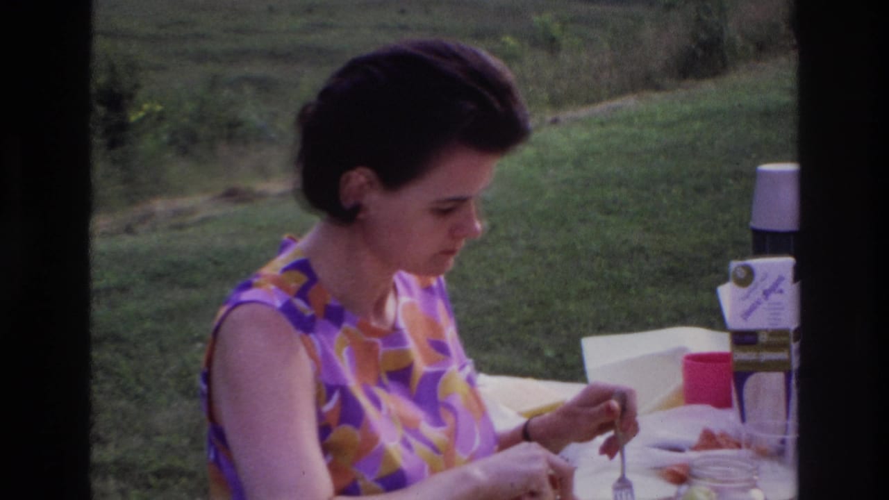 1967 Ottawa home-movie frame showing the woman focused on her meal at an outdoor table with picnic containers visible.