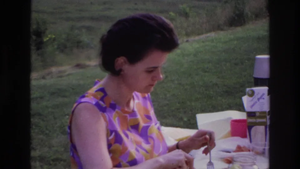 1967 Ottawa home-movie frame showing the woman focused on her meal at an outdoor table with picnic containers visible.