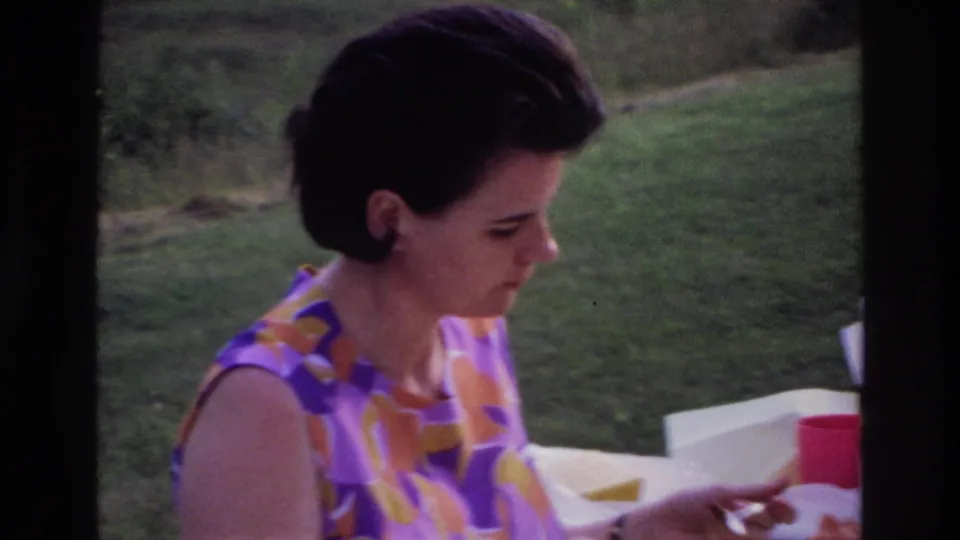 Closer 1967 Ottawa frame of a woman in a floral sleeveless dress eating outdoors against a grassy hillside.