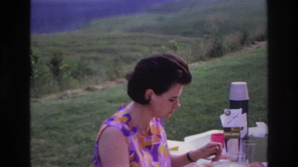 1967 Ottawa home-movie frame of the woman at an outdoor picnic table with a thermos, cup, glass, and hillside behind her.