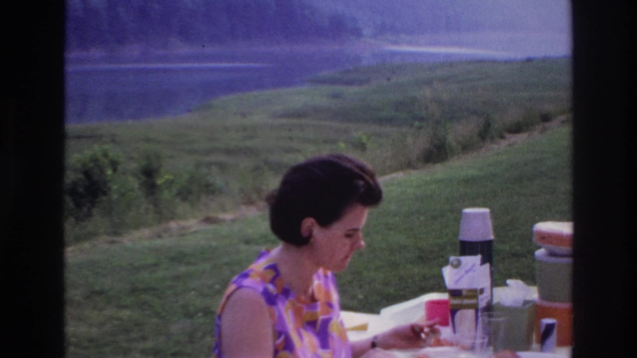 Adult woman in a patterned dress eats at a picnic table in 1967 Ottawa, Canada, with lake and hills behind her.