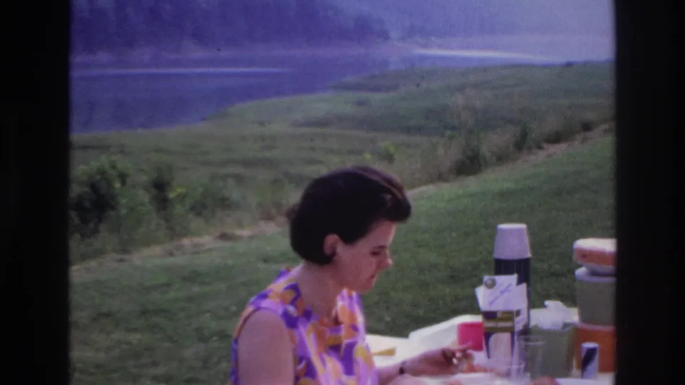 Adult woman in a patterned dress eats at a picnic table in 1967 Ottawa, Canada, with lake and hills behind her.