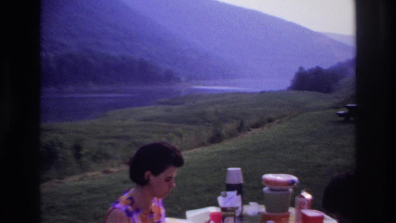 The woman remains at the picnic table as thermos bottles and cups sit in front of a lakeside hill view in 1967 Canada.