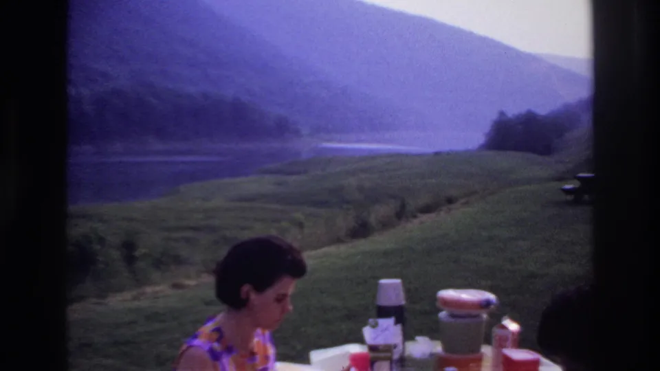 The woman remains at the picnic table as thermos bottles and cups sit in front of a lakeside hill view in 1967 Canada.