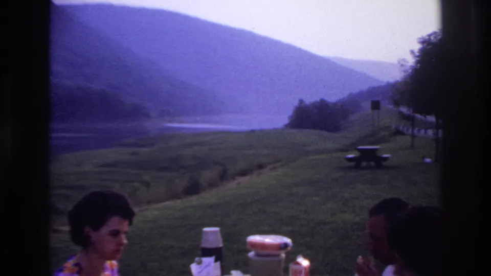 A lakeside picnic scene shows the woman, table items, and a partly visible adult near benches and hazy hills in 1967 Ottawa, Canada.