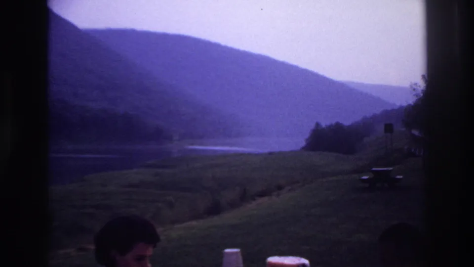 The camera widens to the lakeside hills and picnic grounds, with a partial figure and table items at the bottom edge.