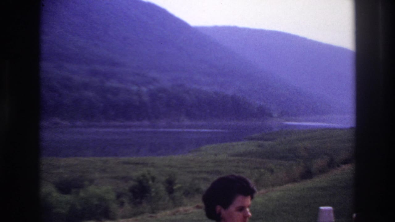 A 1967 home-movie frame shows a calm lake or river below wooded hills, with a partial foreground face near the lower right.
