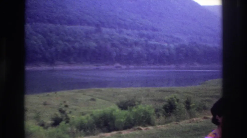 Soft 8mm footage shows a broad Canadian lakeside or riverside view with wooded slopes and a cropped foreground person.