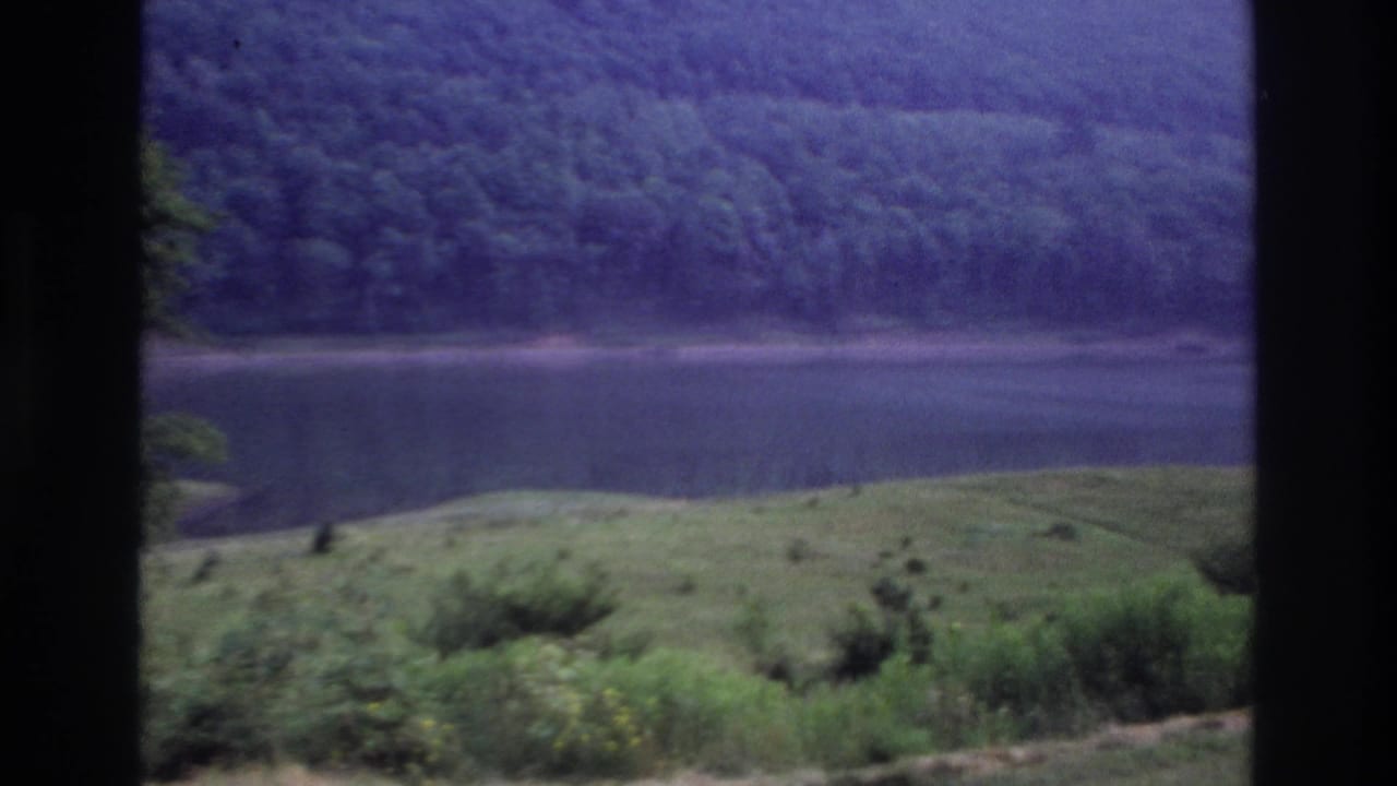 1967 Ottawa home-movie frame showing a calm lake, grassy shore, and wooded hillside through dark vertical framing.