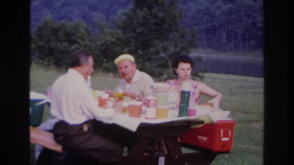 Three adults picnic at a wooden table near a lake in 1967 Ottawa footage, with thermoses, containers, and a red cooler visible.