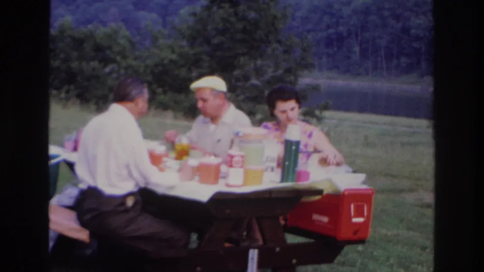 The lakeside picnic continues with three adults at the table, one man in a light cap, and a red cooler at the bench end.