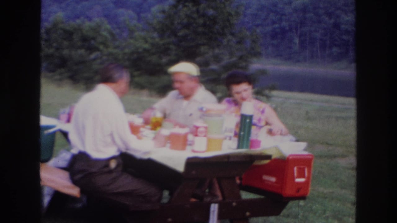 In 1967 Ottawa home-movie footage, three adults sit at a covered picnic table in a grassy area with trees and water behind them.