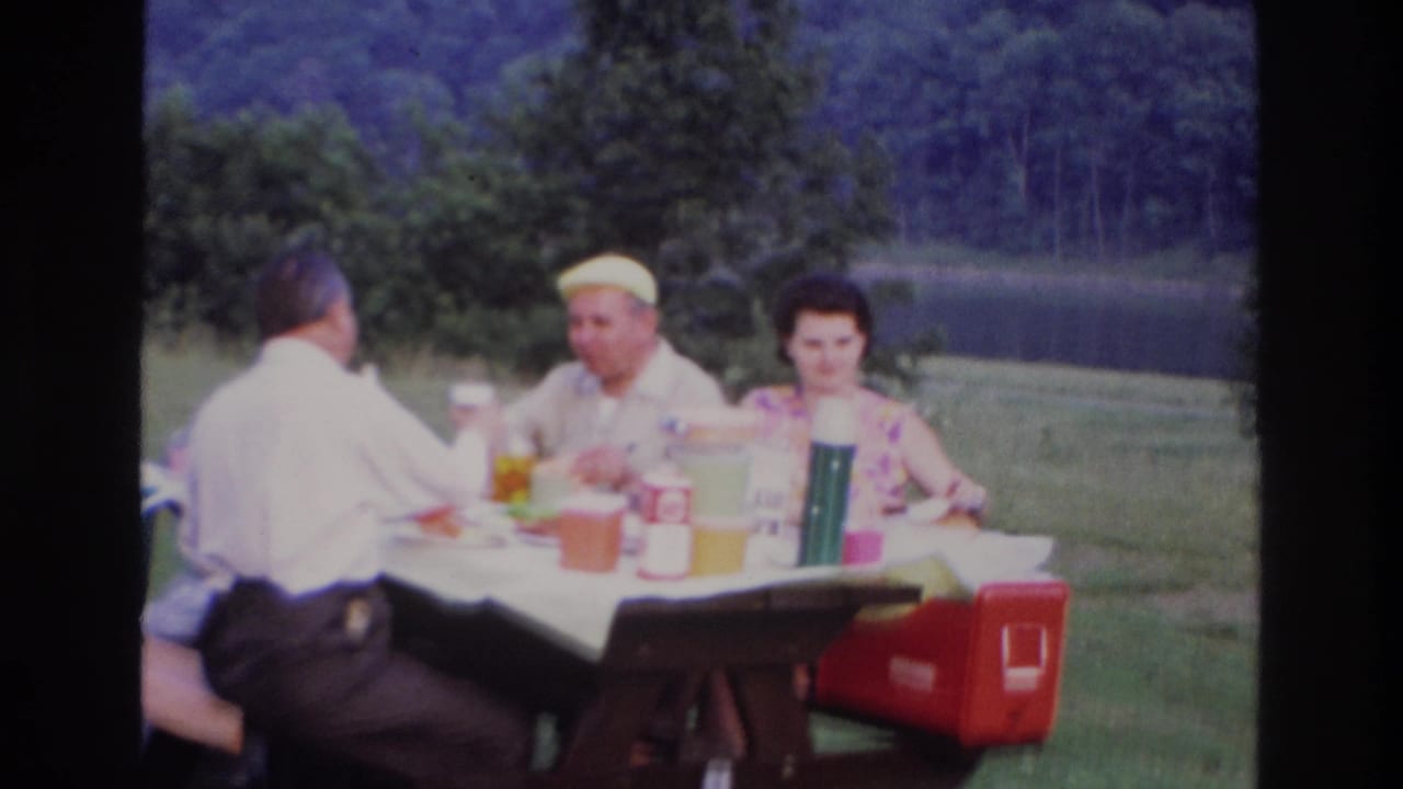 Adults at an outdoor picnic table with a red cooler and drink containers in view.
