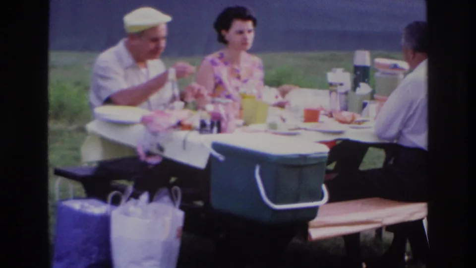 Closer 1967 Ottawa home-movie view of adults eating at a picnic table with coolers and thermoses in the foreground.