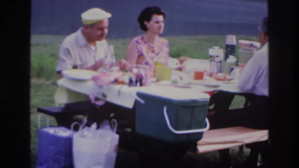 1967 Ottawa-area picnic frame showing adults at a table with portable coolers and a tree-lined body of water beyond.