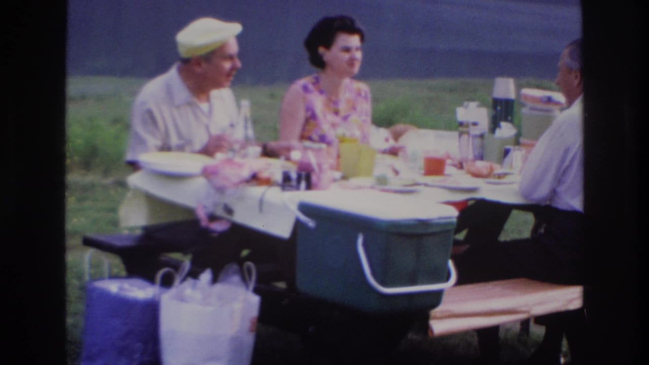 1967 Ottawa home-movie frame of two men and a woman eating and talking at a picnic table in a grassy park.