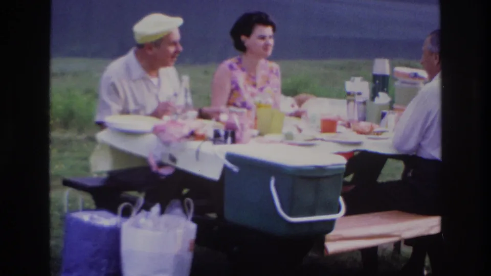 1967 Ottawa home-movie frame of two men and a woman eating and talking at a picnic table in a grassy park.