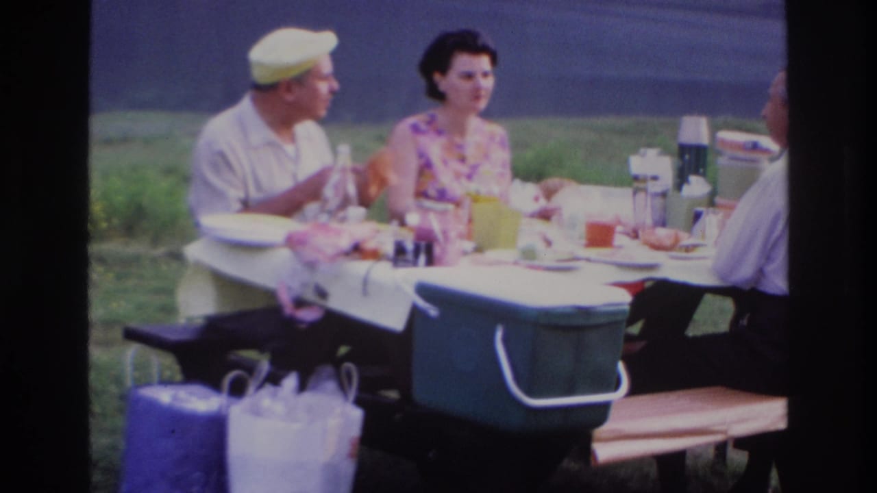 A man in a yellow cap and a woman in a floral dress sit beside a picnic table with food and drinks.