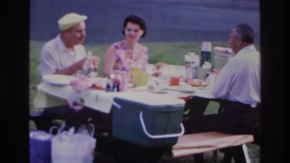 Ottawa 1967 home-movie frame showing three adults conversing over food at a park picnic table.