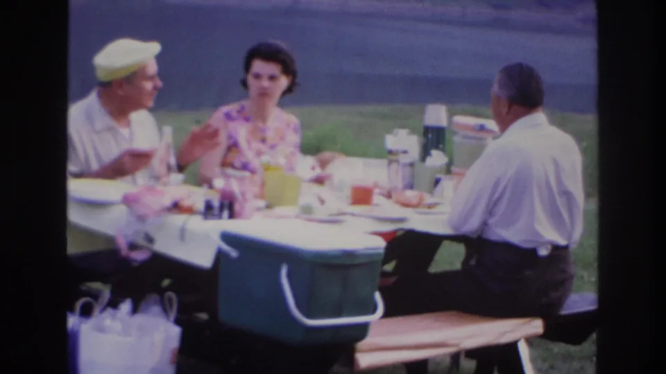 1967 Ottawa park picnic frame with adults seated around a table, food and a green cooler in the foreground.