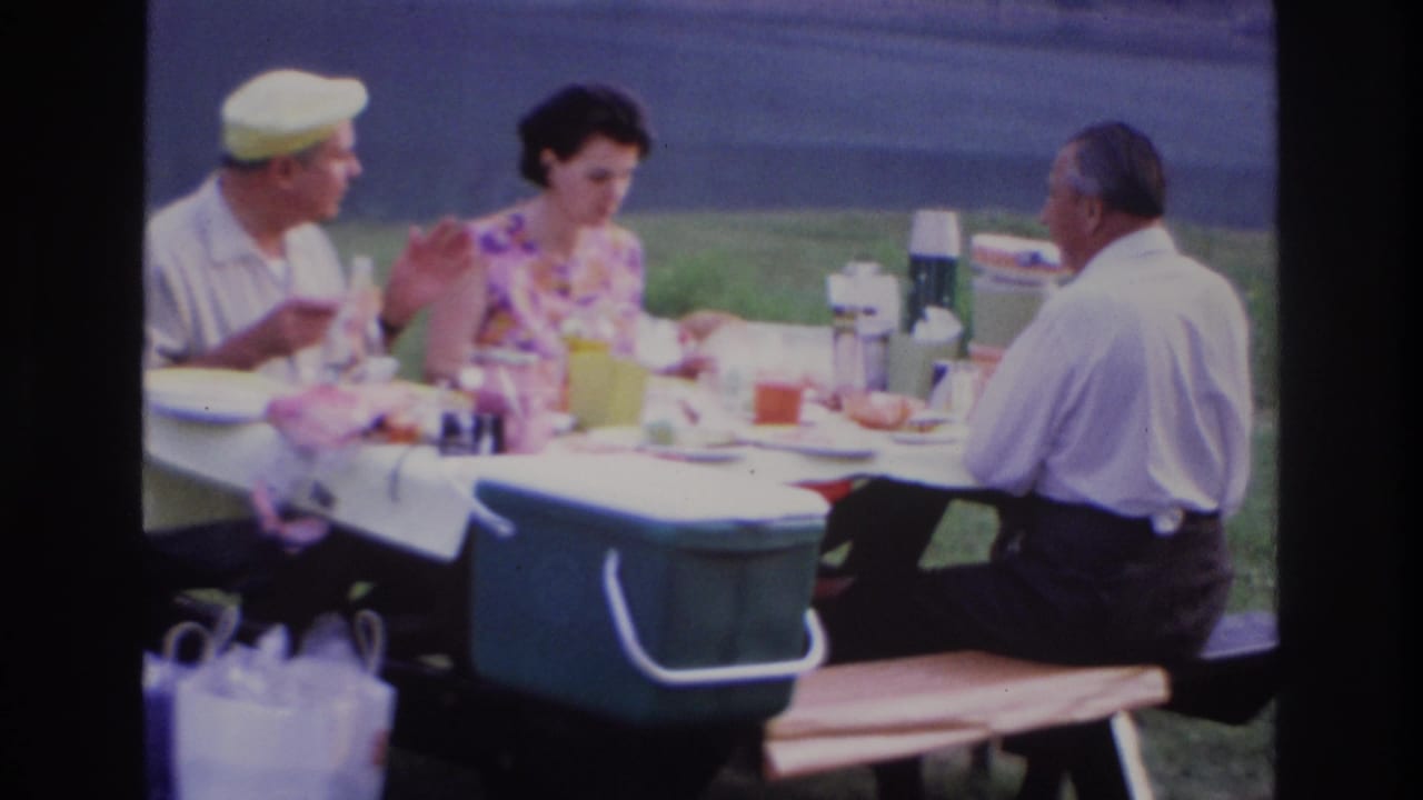 1967 Ottawa home-movie frame showing three adults seated at an outdoor picnic table beside water, with food, thermoses, cups, and a green cooler.