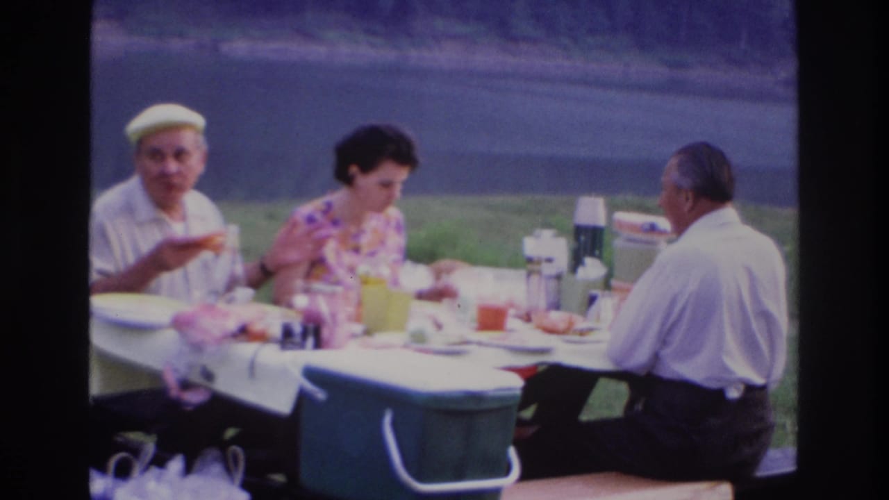 A man in a yellow cap gestures while three adults sit around a picnic table with food and drinks.