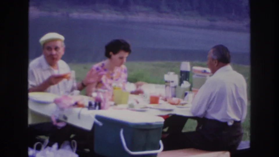 1967 Ottawa picnic frame with a man in a yellow cap, a woman in a floral dress, and another man seated around a table with cups and thermoses.